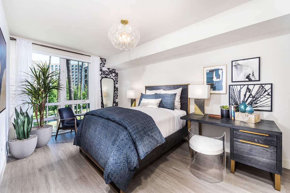 Modernly decorated bedroom at 580 Anton Apartments featuring a spacious bed, desk and chair, nightstands, indoor plants, and a picturesque view of the outside garden.