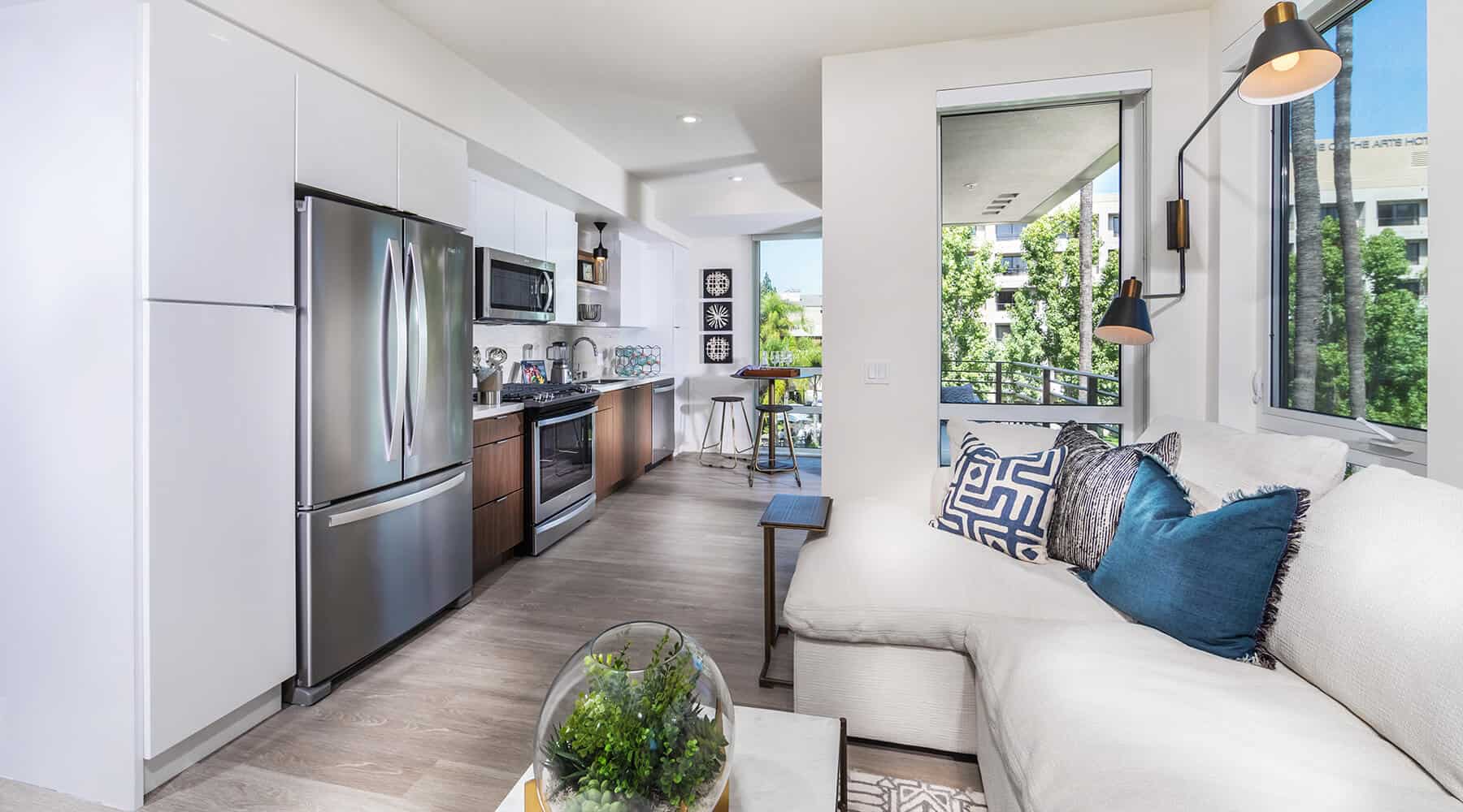 Modern kitchen and living area with a sofa, stainless-steel refrigerator, and large windows with outdoor views.