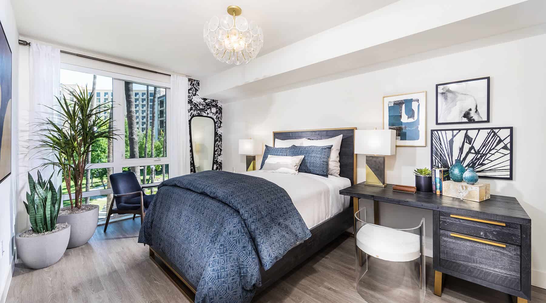 Modernly decorated bedroom at 580 Anton Apartments featuring a spacious bed, desk and chair, nightstands, indoor plants, and a picturesque view of the outside garden.