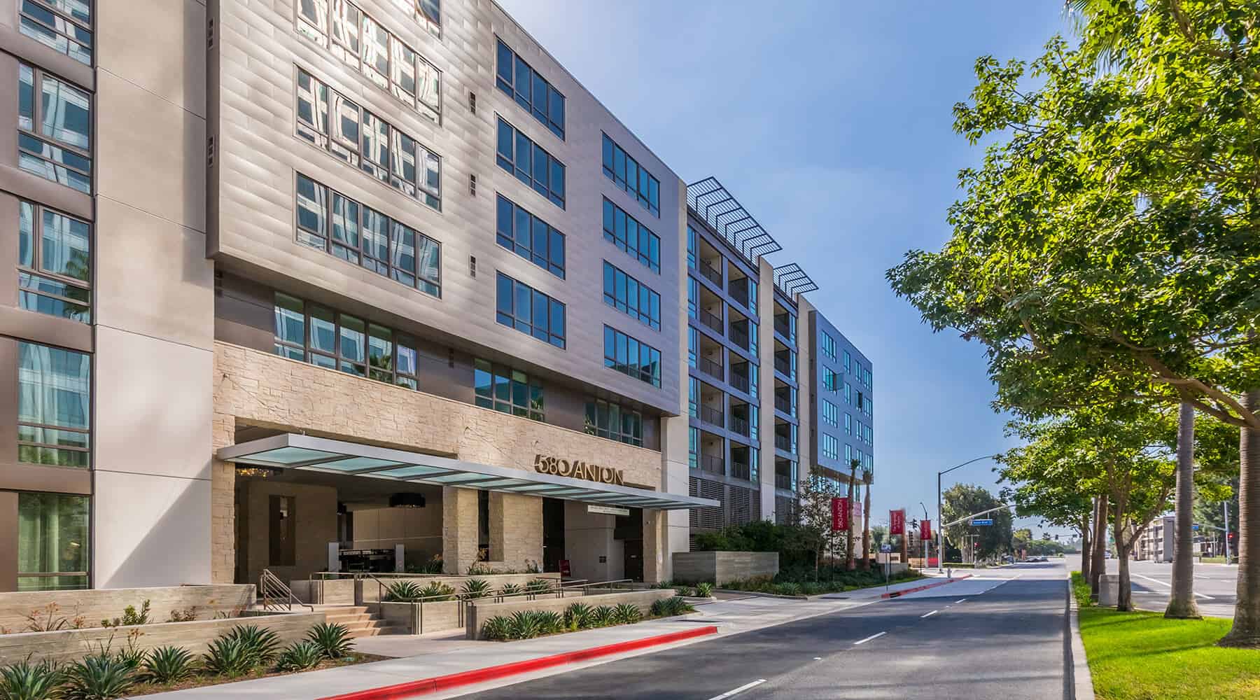 Modern apartment building with glass windows and a covered entrance, surrounded by landscaped greenery.