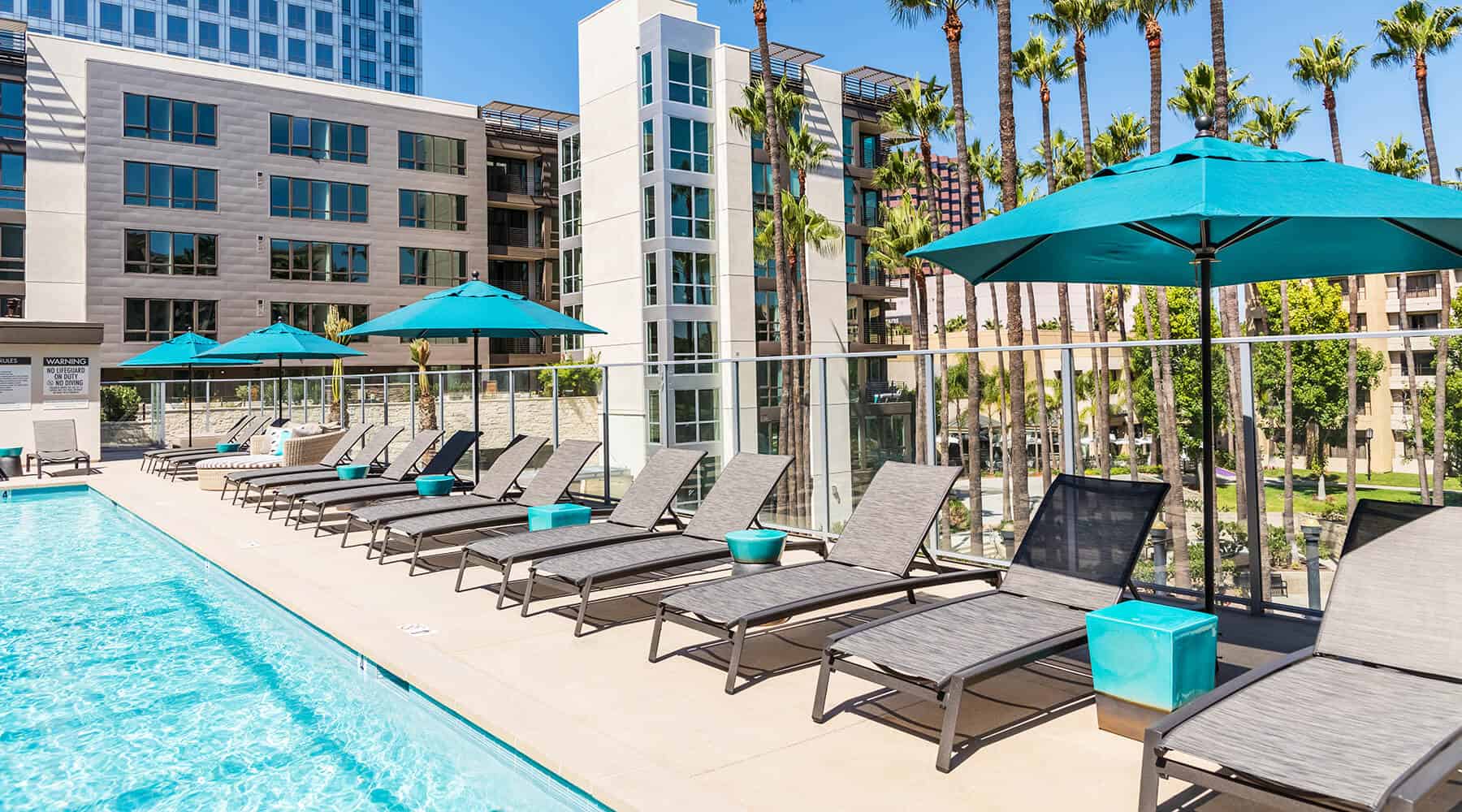 Outdoor pool area with lounge chairs and umbrellas, surrounded by modern apartment buildings and palm trees.