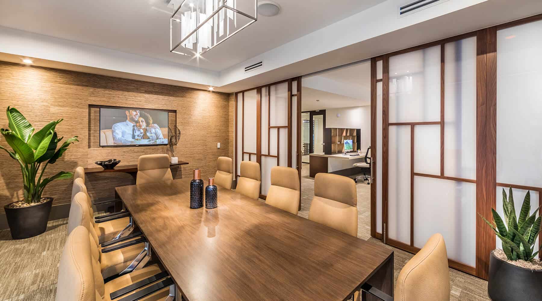 Business meeting room with a large wooden table, comfortable chairs, and a view into an office space with plants and modern decor.